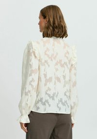 Blusa bianca trasparente con un motivo a foglie testurizzato, dettagli arricciati sulle spalle e maniche gathered. Modello visto di spalle.