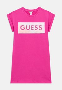 Ροζ βαμβακερό κοντομάνικο μπλουζάκι με ορθογώνιο λευκό πάνελ που φέρει το κείμενο «GUESS» με έντονες ροζ περιγράμματα και διακοσμητικές λεπτομέρειες με καρφίτσες.