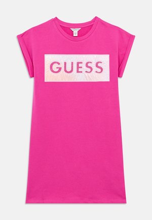 T-shirt rosa in cotone con maniche corte e un pannello rettangolare bianco con la scritta "GUESS" in grassetto, contornata da una linea rosa con dettagli borchiati.