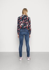 Vuxen som bär en långärmad blommönstrad topp i flera färger, blå slim fit jeans och vita-rosa Nike Air sneakers, stående vänd mot en vit vägg.