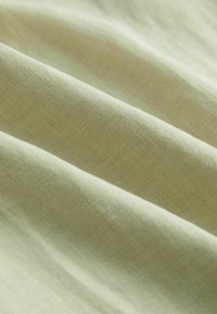 Ljust grönt, texturerat tyg med en fin väv, som visar mjuka veck och subtila variationer i nyans och ljus över ytan.