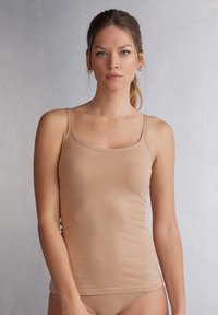 Light beige tank top with thin straps, made from smooth fabric. Features a simple, fitted design and includes a small brand tag at the bottom.