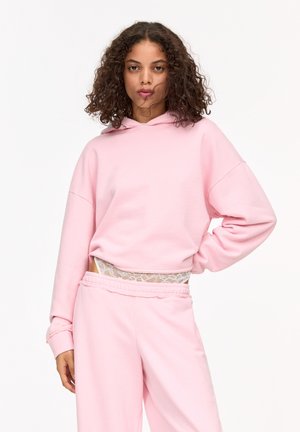 Felpa corta rosa con cappuccio regolabile, tessuto morbido e maniche ampie. Abbinata a pantaloni jogger rosa coordinati con un'accenno di pizzo in vita.