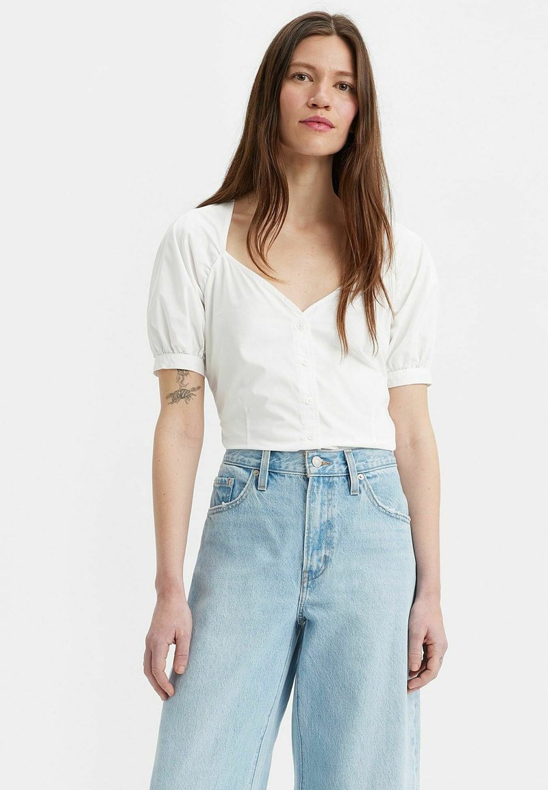 Blusa bianca a maniche corte con scollo a V, chiusura con bottoni e spalle arricciate, abbinata a jeans a gamba larga blu chiaro. Tessuto di cotone liscio.