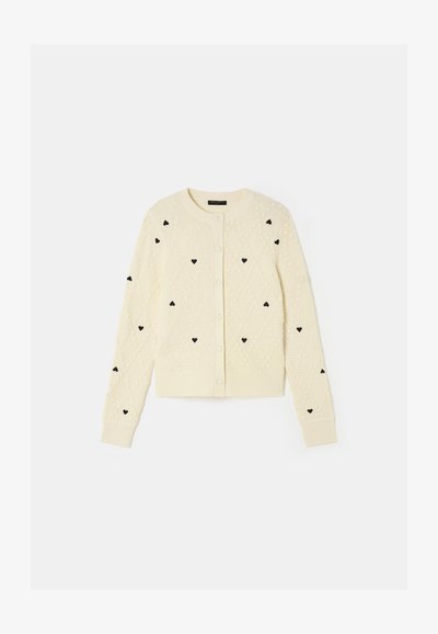 Cardigan crème avec des motifs de cœurs noirs, design en tricot texturé, col rond et fermeture à boutons. Poignets et ourlet côtelés.