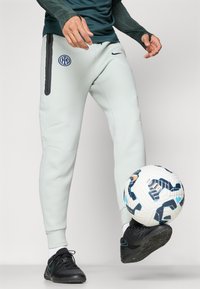 Jasnoszare spodnie sportowe z bocznymi kieszeniami na zamek, z logo Interu Mediolan. W połączeniu z czarnym obuwiem oraz wzorzystą piłką nożną.
