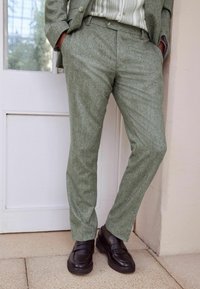 Hellgrüne, strukturierte Anzughosen, slim fit, mit Seitentaschen. Kombiniert mit schwarzen Slippern, vor einem neutralen Hintergrund und einer Tür.