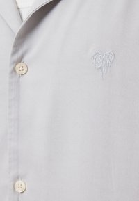 Camicia grigia chiaro con bottoni, caratterizzata da una texture liscia. Presenta un piccolo logo a cuore ricamato in bianco e bottoni beige sul davanti.