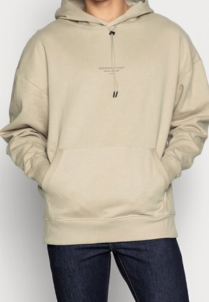Person iført en beige hættetrøje med forlommer og mørkeblå jeans, stående med hænderne i lommerne.