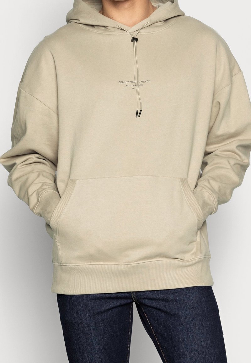 Personne portant un sweat à capuche beige avec des poches avant et un jean bleu foncé, debout, les mains dans les poches.