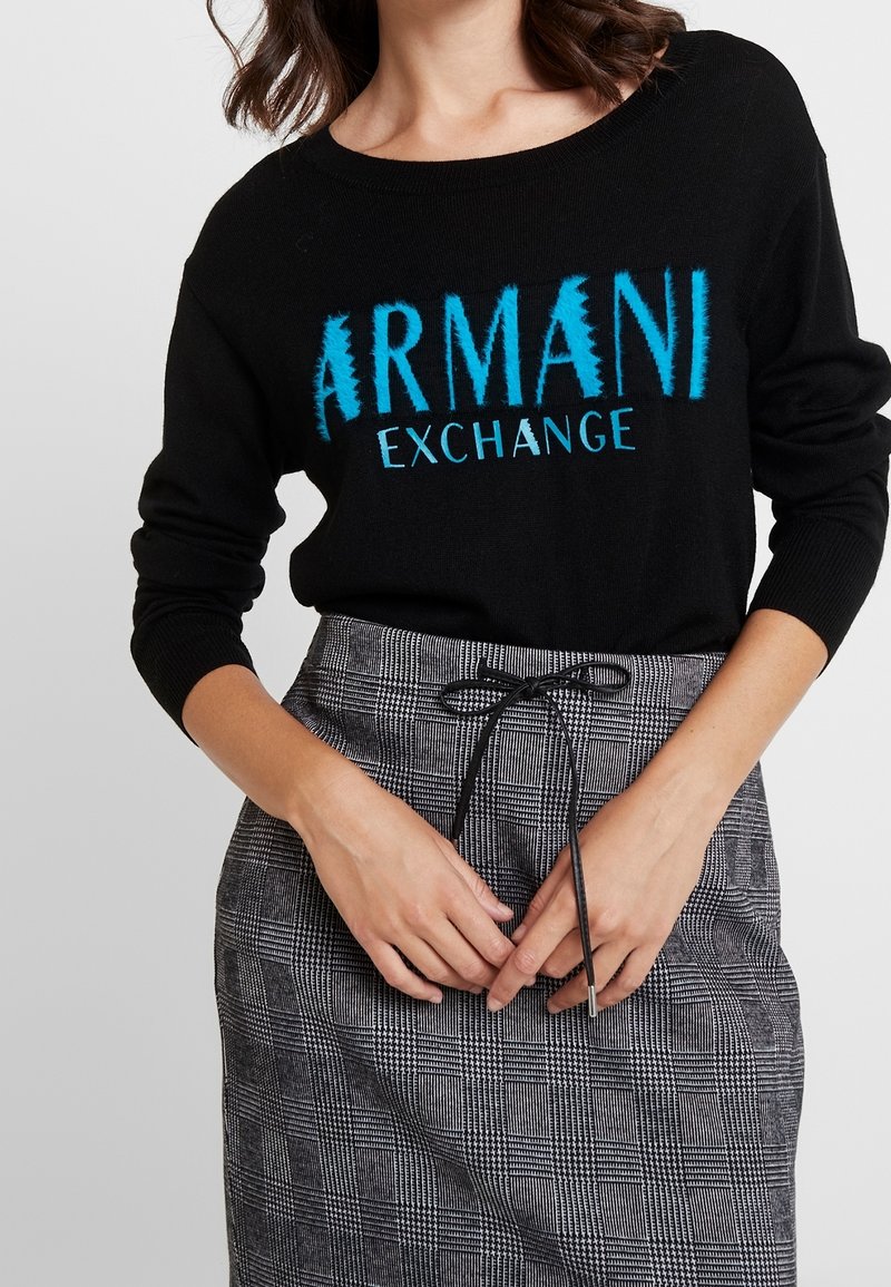 Femme portant un pull noir Armani Exchange avec des lettres bleues, rentré dans une jupe taille haute grise à carreaux, ceinturée d'un cordon noir.