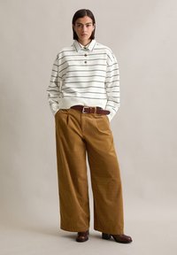 Gestreept wit sweatshirt met houten knopen, gecombineerd met brede bruine corduroybroek. Model draagt bruine haklaarzen en een bruine riem.
