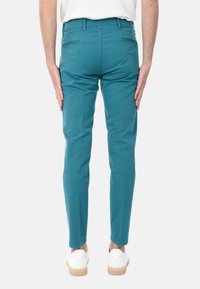 Pantaloni in cotone teal, slim fit, dotati di due tasche posteriori e una texture liscia, abbinati a sneaker bianche su uno sfondo neutro.