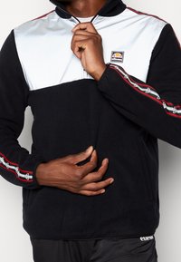 Svart fleece-tröja med en vit nylonövre panel, med en dragkedja vid kragen, röda och svarta accentremsor samt logodetalj på bröstet.