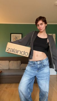 Scatola di cartone con "zalando" stampato in grigio, tenuta da una persona che indossa un top nero corto e jeans blu larghi, in piedi su un pavimento di legno.