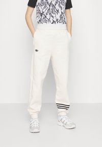 Krémové joggery s elastickým pasem a černými třemi pruhy na manžetách, doplněné vzorovaným pleteným topem a světlými teniskami.