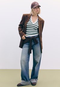 Brun blazer over en grøn og hvid stribet ribbede top, brede udtværede jeans med en knyttet bælte, og brun hat med teksten "Trust the Process".