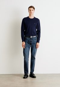Marineblauer Pullover in Strickmuster, kombiniert mit dunkelblauen Jeans, einem schwarzen Gürtel und schwarzen Schuhen. Das Model steht vor einem schlichten weißen Hintergrund.