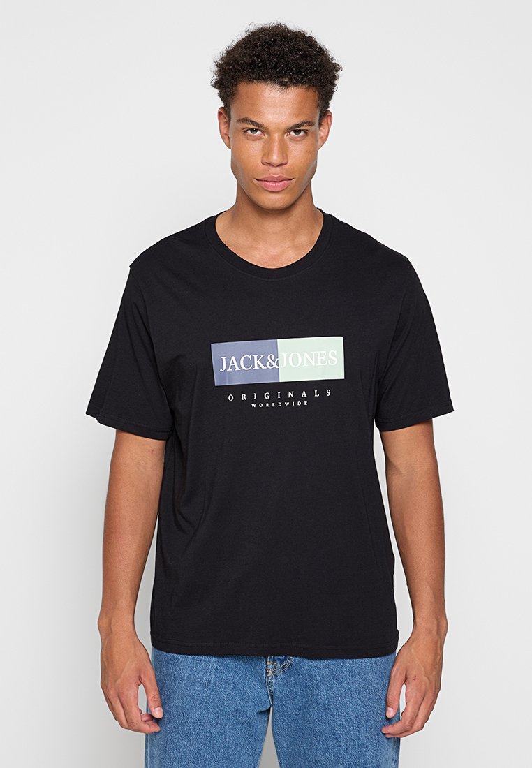 Νέος άνδρας που φοράει μαύρο μπλουζάκι Jack & Jones Originals Worldwide και μπλε τζιν, κοιτάει μπροστά με ουδέτερη έκφραση.