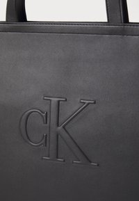 Črna torba iz gladkega materiala z vgraviranim logotipom "CK". Oblika je preprosta z trdnimi ročaji in elegantnim, minimalističnim stilom.