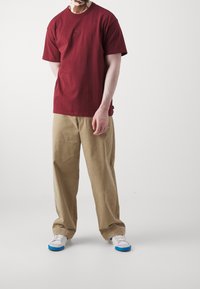 T-shirt di cotone marrone con un piccolo logo, abbinato a pantaloni wide-leg beige e sneakers bianche con accenti blu. Vestibilità casual.