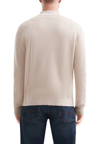 Beige geribbelde gebreide trui met kraag en lange mouwen, heeft een gestructureerde stof en een rechte zoom. Gecombineerd met donkere spijkerbroek.