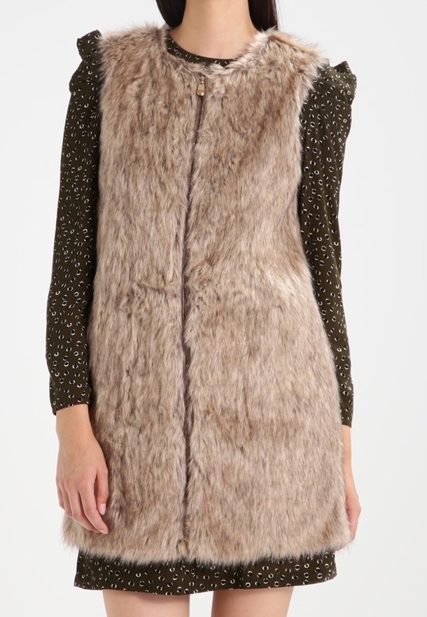 Kamizelka z futerka sztucznego w jasnobrązowym kolorze o miękkiej teksturze, z okrągłym dekoltem i suwakiem z przodu, noszona na wzorzystej sukience w ciemnozielonym kolorze.