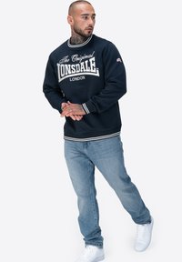 Dunkelblaues Sweatshirt mit weißem Logo, gerippter Hals- und Ärmelbund, kombiniert mit hellblauen Jeans und weißen Sneakers.