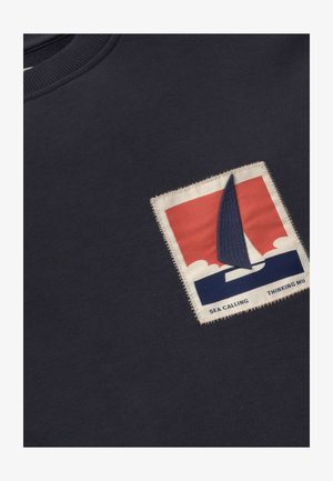 Vêtement bleu marine foncé avec un patch rectangulaire cousu représentant un voilier stylisé sur des vagues rouges et bleues, le texte indique "SEA CALLING THINKING MU".