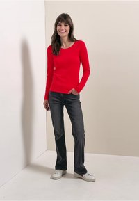 Red long-sleeve top with a scoop neck, paired with dark grey jeans and white sneakers, set against a neutral background.