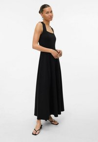 Robe maxi noire sans manches avec un décolleté carré et une silhouette évasée. Tissu lisse, longueur cheville, associée à des tongs noires.