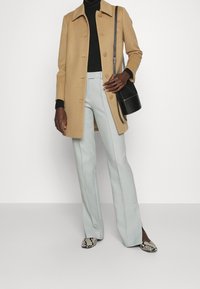 Manteau en laine beige avec un col pointu et des boutons, col roulé noir, pantalon bleu clair ajusté, et chaussures pointues à motifs. Sac à main noir.