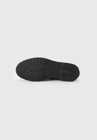 Semelle de chaussure en caoutchouc noir avec des motifs texturés, présentant des rainures et des reliefs pour une meilleure adhérence. Design épuré sans marque ni accents visibles.