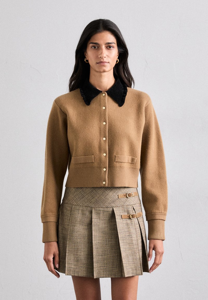 Rudas vilnos megztinis su juodu aksominiu apykakle, mygtukų priekyje ir dviem priekinėmis kišenėmis, derinamas su languota klostuota sijonu su sagtimis.