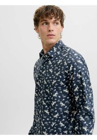 Giovane uomo che guarda a destra, indossando una camicia azzurra a maniche lunghe con un piccolo motivo floreale bianco e beige su uno sfondo chiaro e semplice.