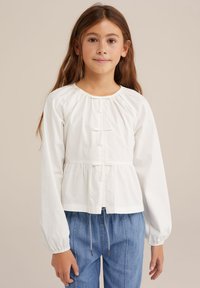 Bluză albă cu mâneci lungi, cu umeri gather și talie peplum. Are un decolteu cu nasturi și este dintr-un material de bumbac moale. Asortată cu blugi albaștri.