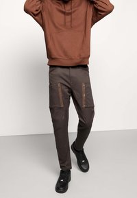 Hnědá mikina s kapsou na přední straně, v kombinaci s tmavě hnědými cargo kalhotami s kapsami na zip. Černé tenisky doplňují celý outfit.