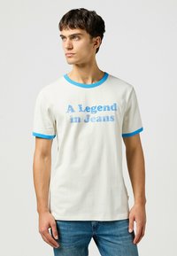Camiseta de algodón blanca con acentos acanalados azules en el cuello y las mangas. Presenta el texto "Una leyenda en vaqueros" en letras azules.