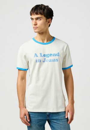 Weißes Baumwoll-T-Shirt mit blauen Rippakzenten am Kragen und an den Ärmeln. Verziert mit dem Text "Eine Legende in Jeans" in blauen Buchstaben.