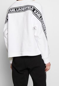 Bílá mikina s ribovaným límcem, dlouhými rukávy a černým grafickým pruhem s nápisem "KARL LAGERFELD" na zadní straně a rukávech.