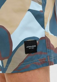 Férfi úszónadrág, amely több színben, absztrakt mintával készült kék, barna és semleges árnyalatokban. A nadrág varrott címkével rendelkezik, rajta a "JACK & JONES EST. 1990." felirattal.