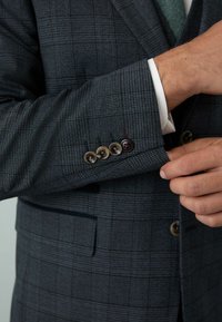 Jachetă de costum în carouri închise, cu un material texturat. Patru butoane pe manșetă în nuanțe diferite. Manșeta cămășii albe care se zărește sub jachetă.