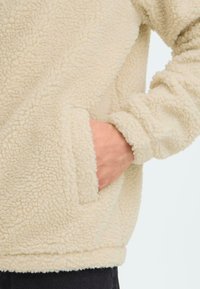 Beige fleece trui met een textuur en een fluffy afwerking en een zijzak. De manchetten en de zoom zijn ook van fleece, wat een zachte, warme uitstraling geeft.
