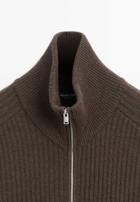 Brązowy sweter z wypustkami, zapinany na zamek z wysokim kołnierzem i srebrnym zamkiem, z charakterystycznym wzorem na tkaninie. Widoczna etykieta marki wewnątrz.
