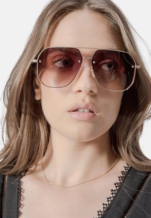 Hexagonale zonnebril met een verloop van donkere roze lenzen, een gouden metalen frame en heldere accenten. Het model draagt een zwart topje met kant en een delicate ketting.