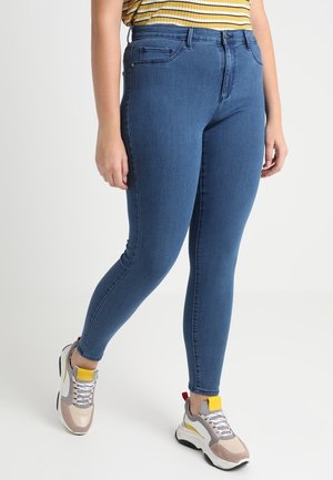 Person iført blå højttaljede skinny jeans, gulstripet top og beige, hvide og gule chunky sneakers, der står på en hvid baggrund.