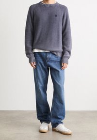 Person trägt einen grauen Rippstrickpullover, bluejeans und weiße Sneaker und steht vor einem schlichten Hintergrund.