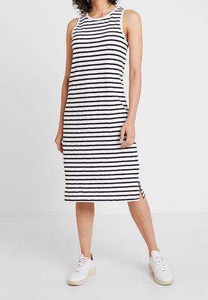 Kobieta w białej, bezrękawnikowej sukience do kolan z czarnymi poziomymi paskami oraz białych tenisówkach, stojąca na jednolitym tle.