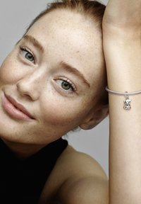 Pulsera de plata con un charm en forma de perro. El modelo tiene pecas claras, ojos verdes y pelo rojo, apoyando su cabeza en la mano. El fondo es gris neutral.
