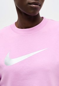 Persoon die een lichtroze T-shirt met ronde hals draagt met een groot wit Nike swoosh-logo op de borst, getoond van nek tot bovenlichaam.